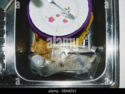 Une vue aérienne de vaisselle sale empilée dans un évier en acier inoxydable. L'assortiment comprend une assiette florale, des contenants en plastique, des ustensiles et des restes Banque D'Images