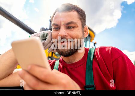 Photographie rapprochée du visage d'un ouvrier agricole utilisant son téléphone intelligent devant la moissonneuse-batteuse Banque D'Images