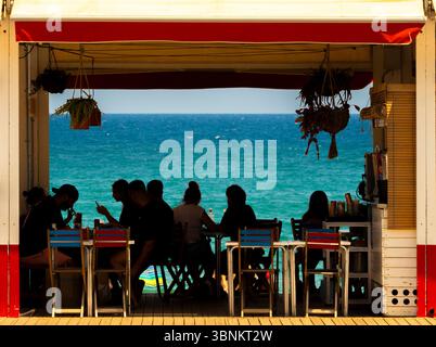 Touristes silhouettés à un bar de plage, ou « chiringuito », à Barcelone, vu dans le bleu vif de la mer Méditerranée en toile de fond. Banque D'Images