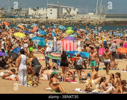 Une plage animée et bondée à Barcelone remplie de gens et de parasols colorés, avec les bateaux et le brise-lames du port visibles en arrière-plan. Banque D'Images