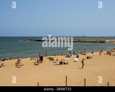 Les amateurs de plage se détendent sur le sable doré de la plage de Somorrostro à Barcelone, avec des eaux méditerranéennes calmes et des navires lointains à l'horizon. Banque D'Images