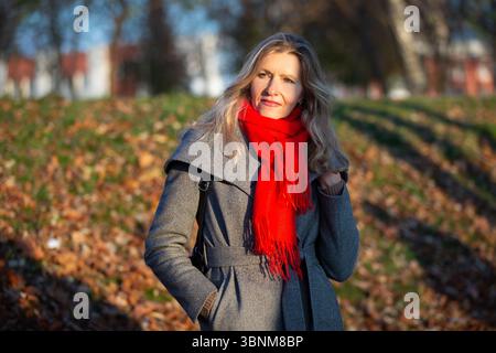 Portrait d'une triste femme blonde de cinquante ans un jour d'automne. Elle porte un manteau gris et une écharpe rouge. Banque D'Images