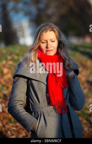Portrait d'une triste femme blonde de cinquante ans un jour d'automne. Elle porte un manteau gris et une écharpe rouge. Banque D'Images