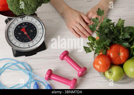 Femme à la table en bois clair avec de la nourriture saine, balance de cuisine, corde à sauter et haltères, vue de dessus Banque D'Images