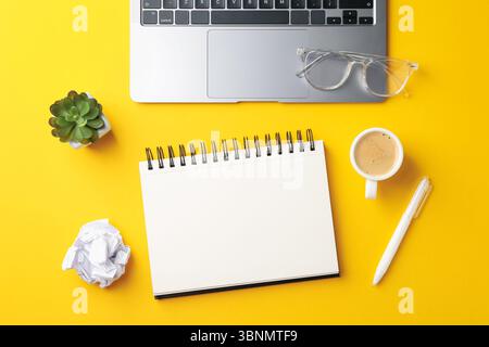 Cahier, stylo, verres, café, ordinateur portable et décor floral sur fond jaune, plat Banque D'Images