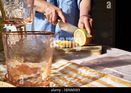 Gros plan d'une femme tranchant un citron sur une planche de bois. La lumière du soleil met en valeur les agrumes frais, la verrerie, et une ambiance estivale confortable parfaite pour la limonade ou un b Banque D'Images