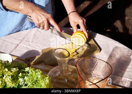 Gros plan d'une femme tranchant un citron sur une planche de bois. La lumière du soleil met en valeur les agrumes frais, la verrerie, et une ambiance estivale confortable parfaite pour la limonade ou un b Banque D'Images