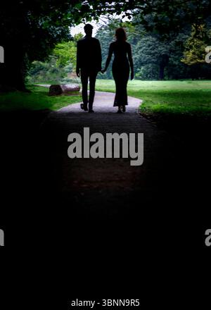 Un couple marche main dans la main sur un chemin dans un parc, silhouetté à contre-courant. Banque D'Images