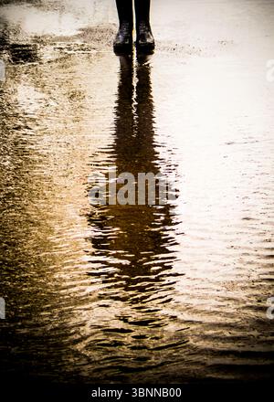 Une personne portant des bottes noires se tient dans une flaque, leur reflet visible dans l'eau. Banque D'Images