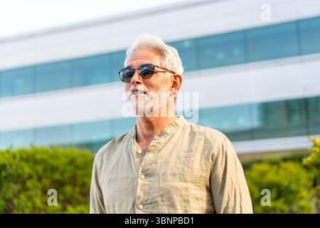 Homme d'affaires mûr avec les cheveux gris et la barbe portant des lunettes de soleil debout avec confiance près de l'immeuble de bureaux en verre moderne avec de la verdure en arrière-plan Banque D'Images
