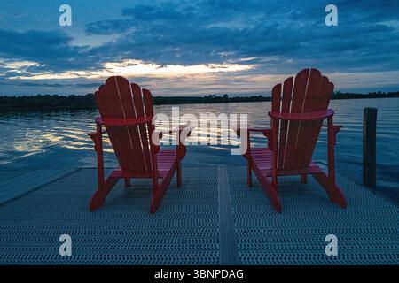 Deux chaises Adirondack rouges au bord d'un quai sur la baie Georgienne dans le lac Huron en Ontario, Canada au coucher du soleil avec des nuages dans le ciel. Banque D'Images