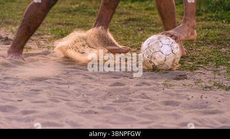 Joueurs pieds nus participant à un match de football animé sur une plage de sable, avec la poussière et les mouvements capturés dans les moindres détails. Banque D'Images