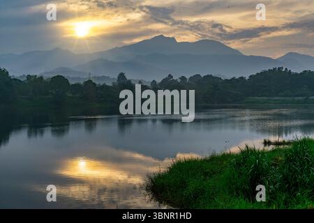 Lever de soleil sur le lac calme avec des reflets de montagne, ciel brumeux, et verdure luxuriante dans un paysage naturel paisible. Banque D'Images