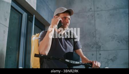 Livreur sur scooter électrique debout près du bureau, acceptant une nouvelle commande du client parlant par téléphone. Homme travaillant dans le service de livraison de repas, livrant la nourriture du supermarché, restaurant. Ralenti Banque D'Images