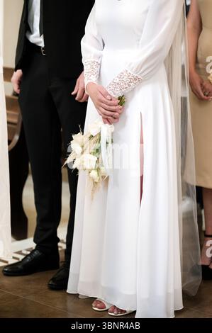 Une mariée se tient dans une église, tenant un petit bouquet. Elle porte une robe blanche fluide avec une fente haute, et un voile. Un homme en costume noir se tient debout Banque D'Images