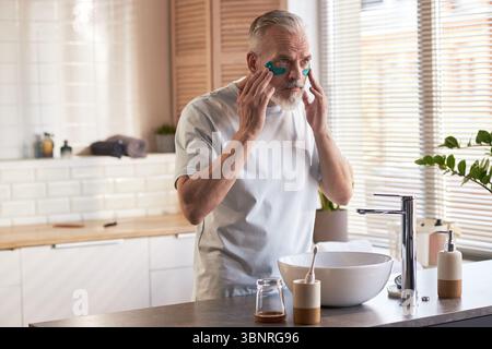 Homme caucasien d'âge moyen appliquant sous les patchs oculaires tout en se tenant debout à l'évier de salle de bain, en se concentrant sur la routine de soins de la peau, les mains pressant doucement les patchs sur le visage, la lumière naturelle illuminant la scène Banque D'Images