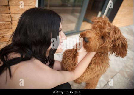 Une femme aux cheveux longs et sombres sourit alors qu'elle tient et embrasse son petit Goldendoodle brun et moelleux. Ils se tiennent près d'un mur de briques et d'une fenêtre, baignés dedans Banque D'Images