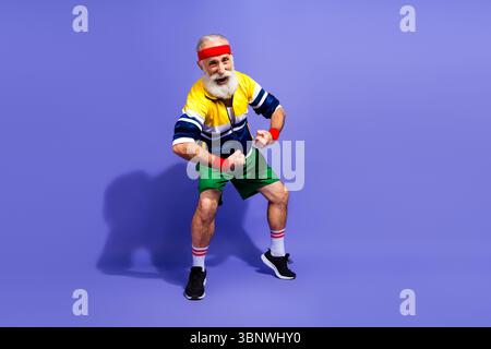 Homme senior énergique dans les vêtements de sport rétro posant avec confiance sur fond violet vibrant exprimant le bien-être et le style de vie de fitness Banque D'Images