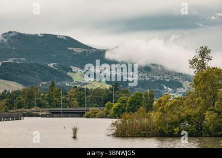 Paysage pittoresque de la campagne suisse avec des collines boisées, un lac au premier plan, et un village brumeux niché parmi les pentes verdoyantes. Capturé dans Banque D'Images
