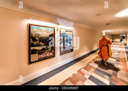 L'Hotel Kempinski Engelberg, un moine bouddhiste vêtu de robes orange, descend le long du couloir de l'hôtel devant des affiches publicitaires historiques pour les vacances à Engelberg. Un moine bouddhiste traverse un couloir de l'hôtel Kempinski à Engelberg, Obwald, Suisse. Les murs sont ornés d'affiches de voyage vintage faisant la promotion de la région d'Engelberg et du chemin de fer d'Engelberg Banque D'Images