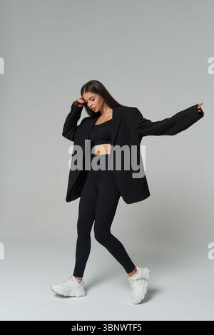 Une femme portant un soutien-gorge de sport noir et des leggings, associée à un blazer noir surdimensionné et des baskets blanches, prend une pose gracieuse. Ses longs cheveux bruns Banque D'Images