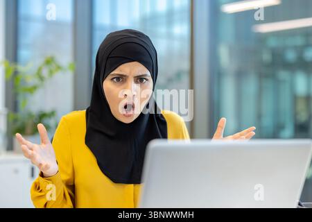 Une musulmane étonnée réagit avec surprise en regardant son ordinateur portable dans un bureau. Banque D'Images