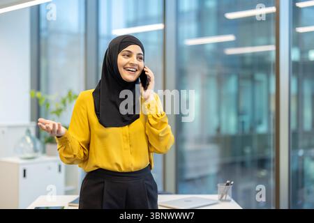 Une femme joyeuse portant un hijab parle au téléphone dans un bureau lumineux. Banque D'Images