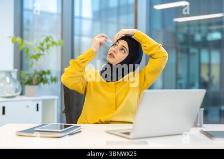 Une femme d'affaires portant un hijab applique des gouttes pour les yeux dans un environnement de bureau moderne, susceptibles de soulager la fatigue oculaire tout en travaillant sur un ordinateur portable. Banque D'Images