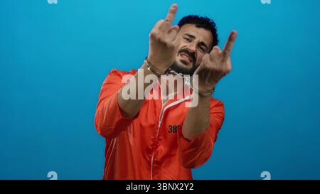 Homme hispanique en combinaison prison orange avec menottes pose sur fond bleu véhiculant une scène dramatique d'incarcération et de défiance. Banque D'Images