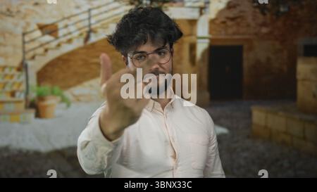 Jeune homme barbu à l'extérieur sur une journée ensoleillée faisant un geste grossier montrant le majeur dans la cour avec des murs de pierre et escalier en arrière-plan Banque D'Images