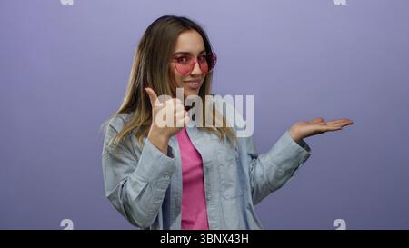 Femme faisant un geste dans une chemise bleue avec une expression ludique pointant vers sa paume ouverte sur un fond violet vif portant des lunettes de soleil roses et Banque D'Images