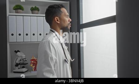Jeune homme portant un manteau blanc et un stéthoscope regardant par la fenêtre dans la salle de clinique moderne avec des dossiers et un microscope à proximité. Banque D'Images