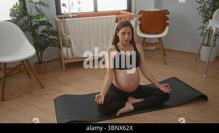 Femme enceinte faisant du yoga sur un tapis dans une chambre avec un berceau à côté d'elle, mettant en valeur une atmosphère sereine et se concentrant sur l'exercice prénatal. Banque D'Images