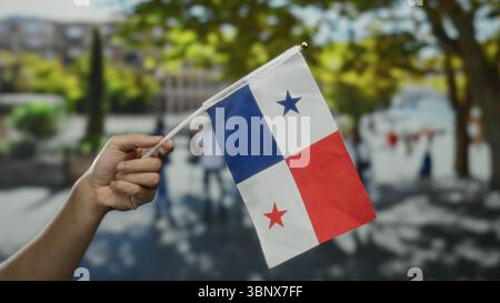 Main de l'homme caucasien tenant le drapeau du panama sur la rue de la ville avec des gens flous et des arbres en arrière-plan, symbolisant la fierté nationale et l'outdo de célébration Banque D'Images