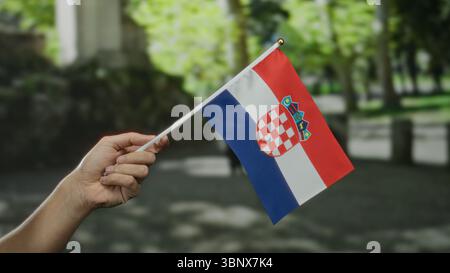 Main tenant le drapeau croate à l'extérieur dans la rue de la ville avec de la verdure, représentant la fierté nationale, l'identité et la célébration dans un cadre extérieur vibrant. Banque D'Images