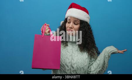 Femme portant le chapeau de père noël et le pull tient le sac à provisions rose avec l'expression confuse devant le fond bleu indiquant le dilemme du shopping de vacances. Banque D'Images