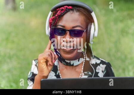 Jeune femme noire confiante avec des lunettes écoutant de la musique avec des écouteurs à l'extérieur tout en utilisant un ordinateur portable. Parfait pour le streaming audio, podcast, vie Banque D'Images