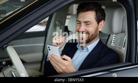 Excité homme d'affaires caucasien voiture heureux en utilisant le téléphone mobile smartphone réalisation Guy célébrer le succès automobile pilote adulte gagner acheteur bon Banque D'Images