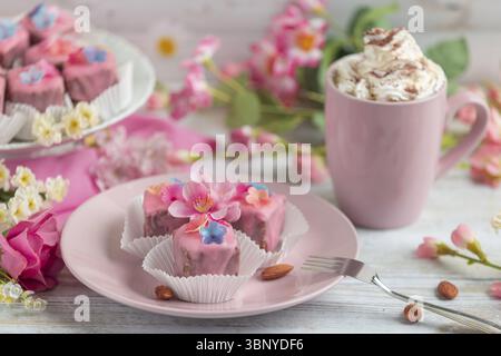 Petits fours roses avec décoration florale sur une assiette à côté d'une tasse de café à la crème Banque D'Images