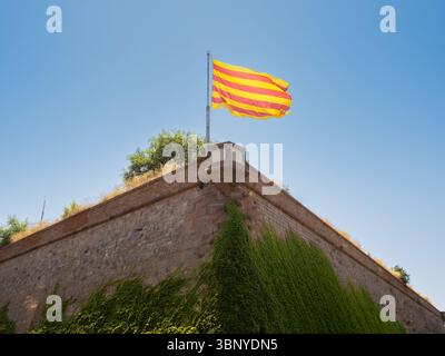 Le drapeau catalan, la Senyera, flotte fièrement des remparts en pierre historiques du château de Montjuïc à Barcelone, en Espagne, par temps clair. Banque D'Images