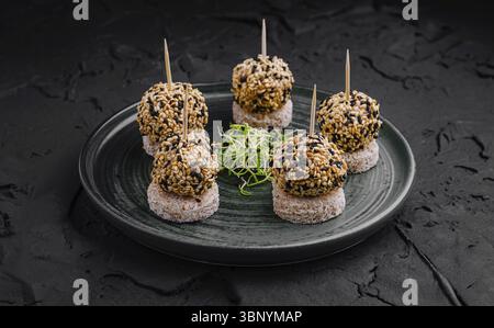 Natures mortes, six boules enrobées de sésame sont disposées sur un plateau sombre, reposant sur de petites croûtes de pain. Les verts frais sont présentés avec art, enhanc Banque D'Images