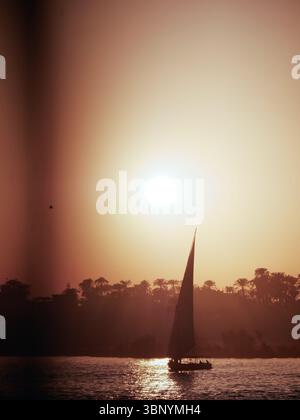 Une silhouette de voilier glisse gracieusement sur une rivière tranquille alors que le soleil se couche en Égypte, peignant le ciel avec des teintes chaudes. Banque D'Images