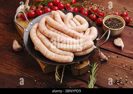 Nature morte, saucisses sont exposées sur une poêle en fonte, entourées de tomates cerises fraîches, d'ail et de diverses épices sur une surface en bois, c Banque D'Images