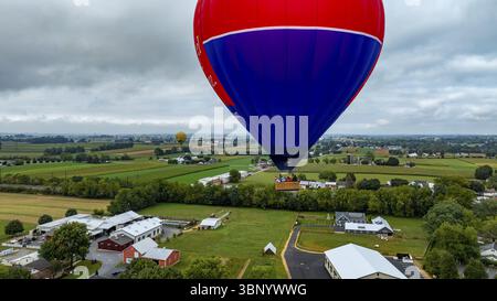 Une montgolfière vibrante survole de vastes champs verdoyants et de structures agricoles sous un ciel nuageux. Le cadre rural offre une toile de fond sereine Banque D'Images