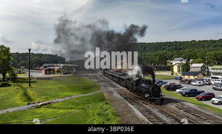 Une locomotive à vapeur longe les voies ferrées, dégageant des nuages de fumée sur fond de paysages verdoyants et d'une ancienne gare. Le paramètre Banque D'Images
