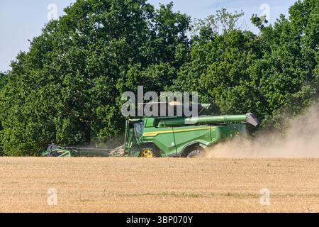 4 juillet 2025 - Lauterbach-Allemagne : une moissonneuse-batteuse John Deere travaille dans un champ d'orge, soulevant la poussière lorsqu'elle récolte une pente avec des arbres à l'intérieur Banque D'Images
