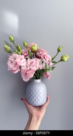 Beau bouquet de fleurs en bourgeonnement d'œillets roses doux dans un vase blanc délicat tenu sur un fond gris doux Banque D'Images