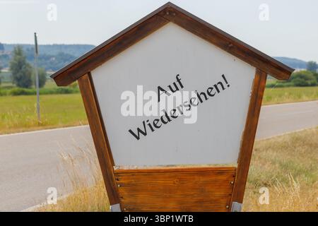 Un panneau en bois avec 'Auf Wiedersehen !' en noir, le texte se tient près d'une route contre un fond herbeux et rural sous un ciel lumineux. Banque D'Images