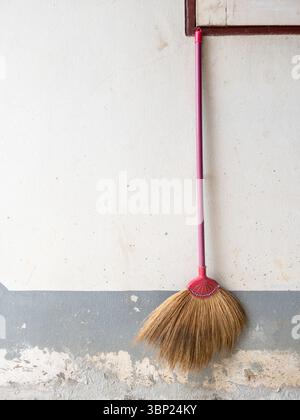 Un vieux balai avec un manche en bois et des poils de paille accrochés sur un mur de maison rurale fané, soulignant le stockage traditionnel des outils de nettoyage Banque D'Images