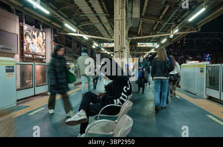 Au quai d'une gare de Tokyo, une personne portant un sweat à capuche repose sur un banc tandis que les navetteurs passent Banque D'Images